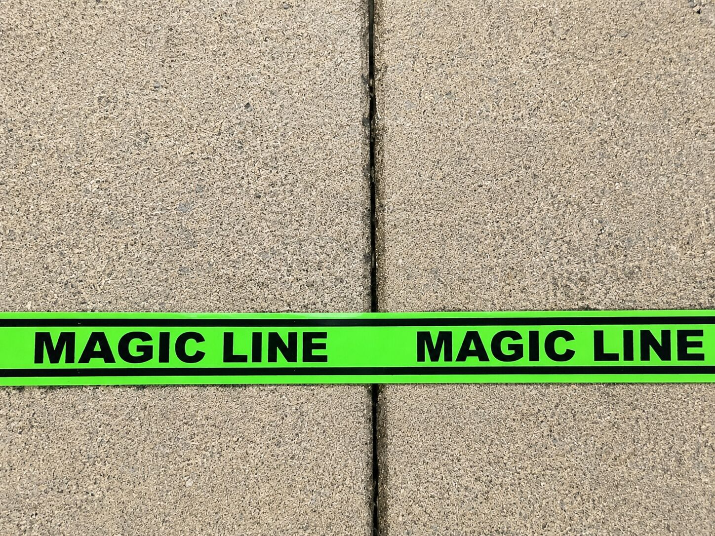 A strip of fluorescent green Magic Line tape with black lettering, applied straight across grey paving slabs.